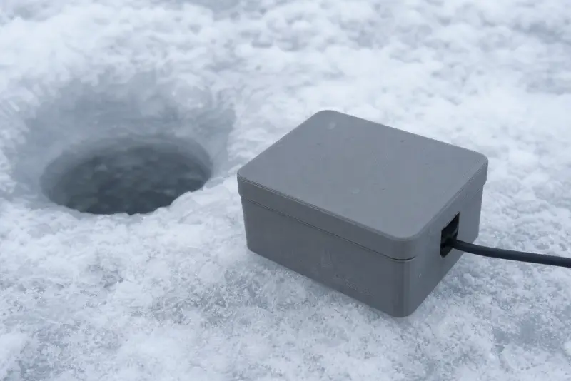 Grå 3D-printet boks med lokk og kabel ved et isfiskehull på et snødekt vann
