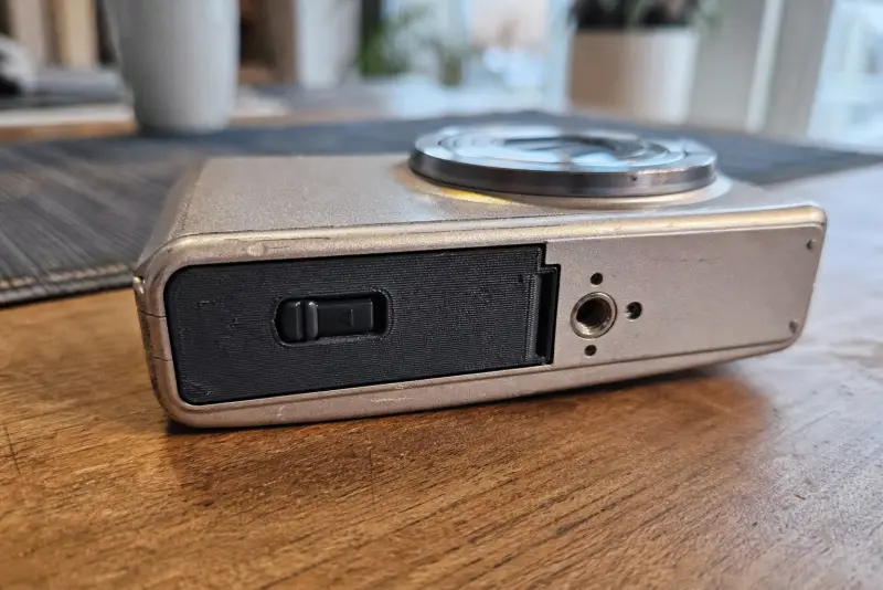 Nytt svart 3D-printet batterilokk på et eldre kompaktkamera på et trebord