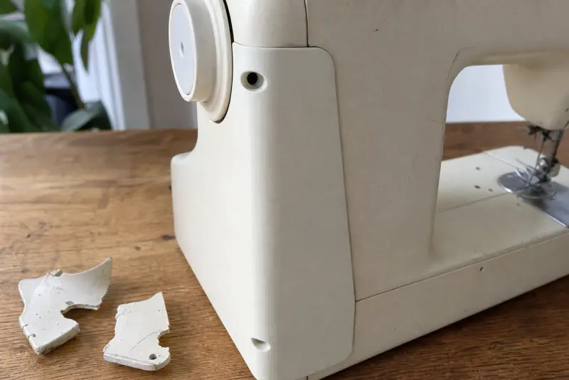 Nytt offwhite 3D-printet deksel på siden av en gammel symaskin, bitene av det gamle dekselet ved siden av
