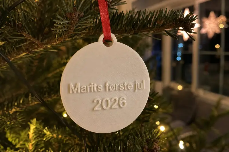 Flat hvit 3D-printet julepynt med teksten Marits første jul 2026 på et juletre