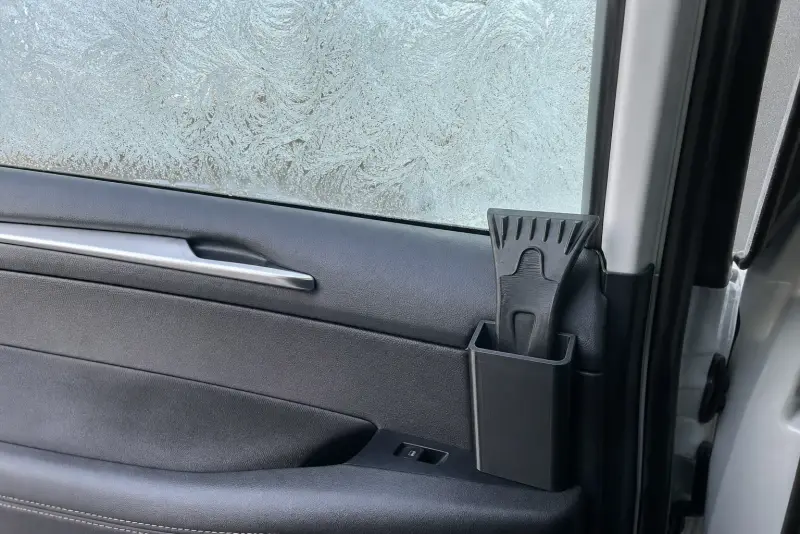 Svart 3D-printet holder med en isskrape i dørlomma på en bil med rim på ruta