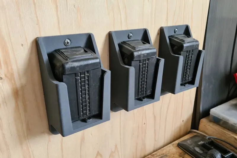 Tre grå 3D-printede veggholdere for verktøybatterier på en kryssfinervegg