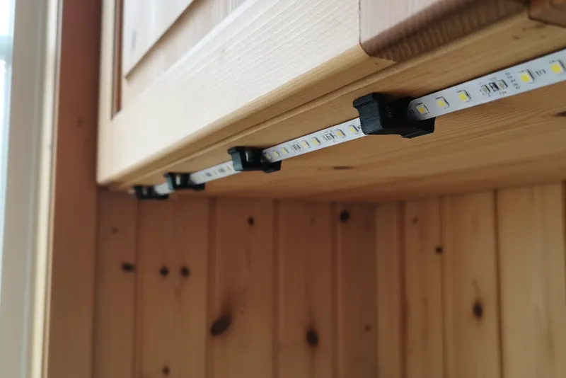 Små svarte 3D-printede klips som holder en tynn LED-list under et kjøkkenskap i tre på en hytte