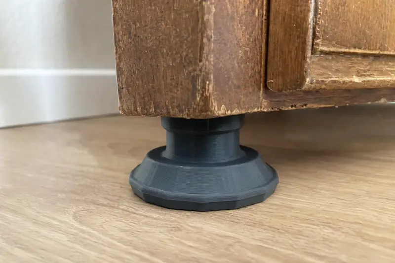 Ny mørkegrå 3D-printet fot under beinet på et gammelt treskap på et lyst eikegulv