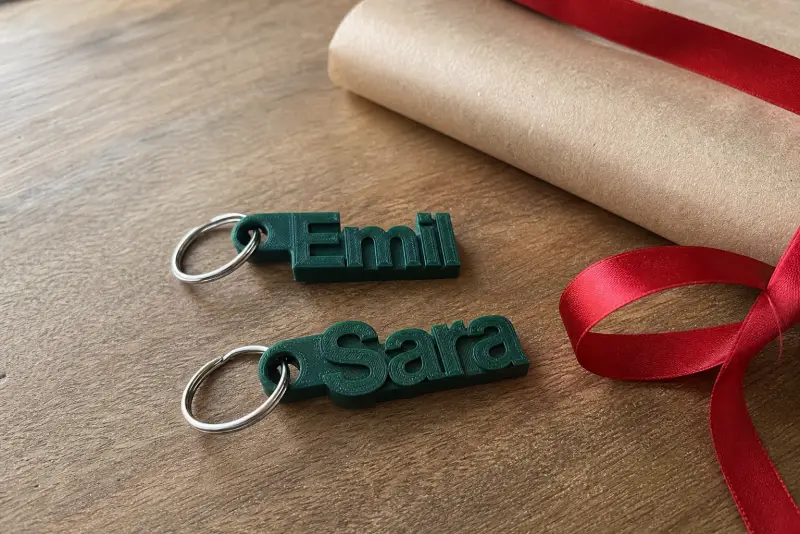 Grønne 3D-printede nøkkelringer med navnene Emil og Sara på et bord ved siden av gavepapir