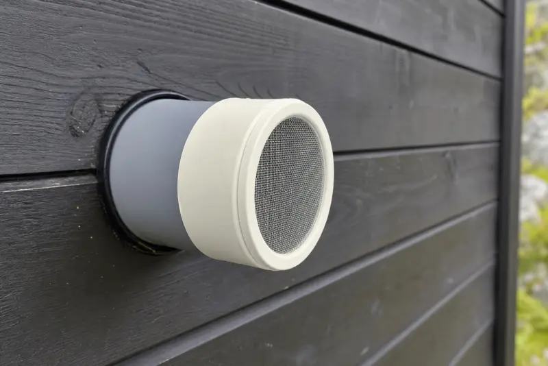 Offwhite 3D-printet rørende med fint insektnett på et ventilasjonsrør i en malt hyttevegg