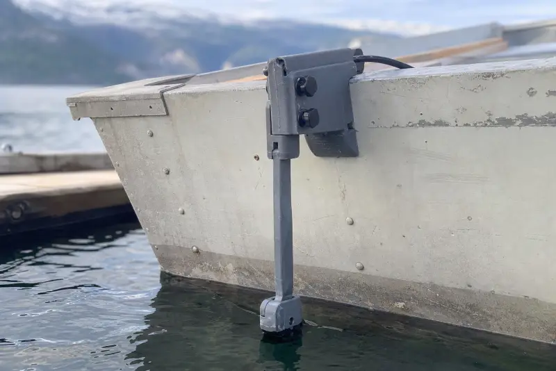 Grå 3D-printet svingerarm klemt fast på akterspeilet til en gammel aluminiumsrobåt ved en brygge