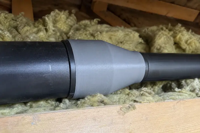 Grå 3D-printet overgang mellom et tykt og et tynt ventilasjonsrør på et hytteloft med isolasjon